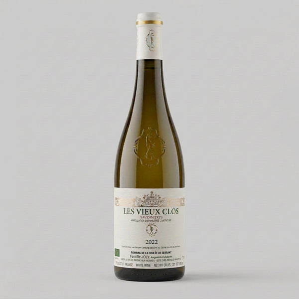 A bottle of Les Vieux Clos 2022 red wine in a dark glass bottle with a cream-colored label and gold foil, set on a wooden surface with a blurred wine cellar background.