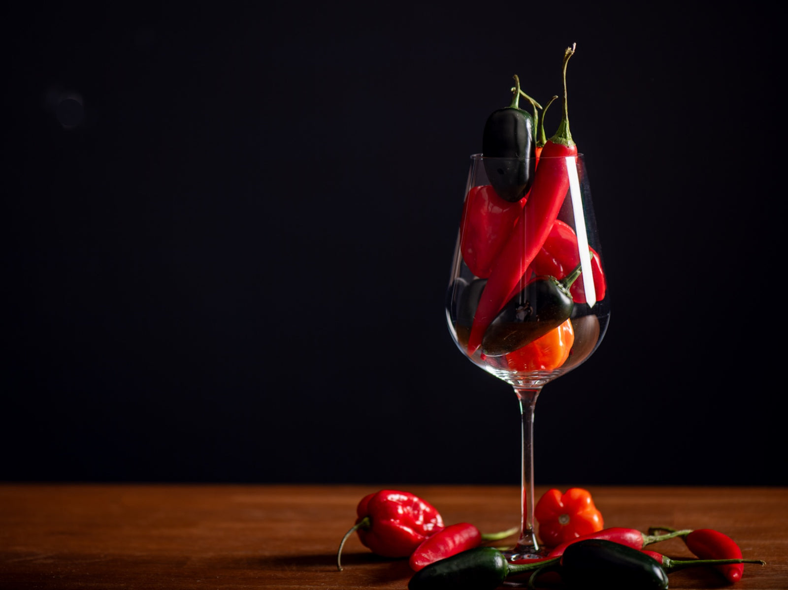 A wine glass filled with red and green chillies, with more chillies on a wooden table.